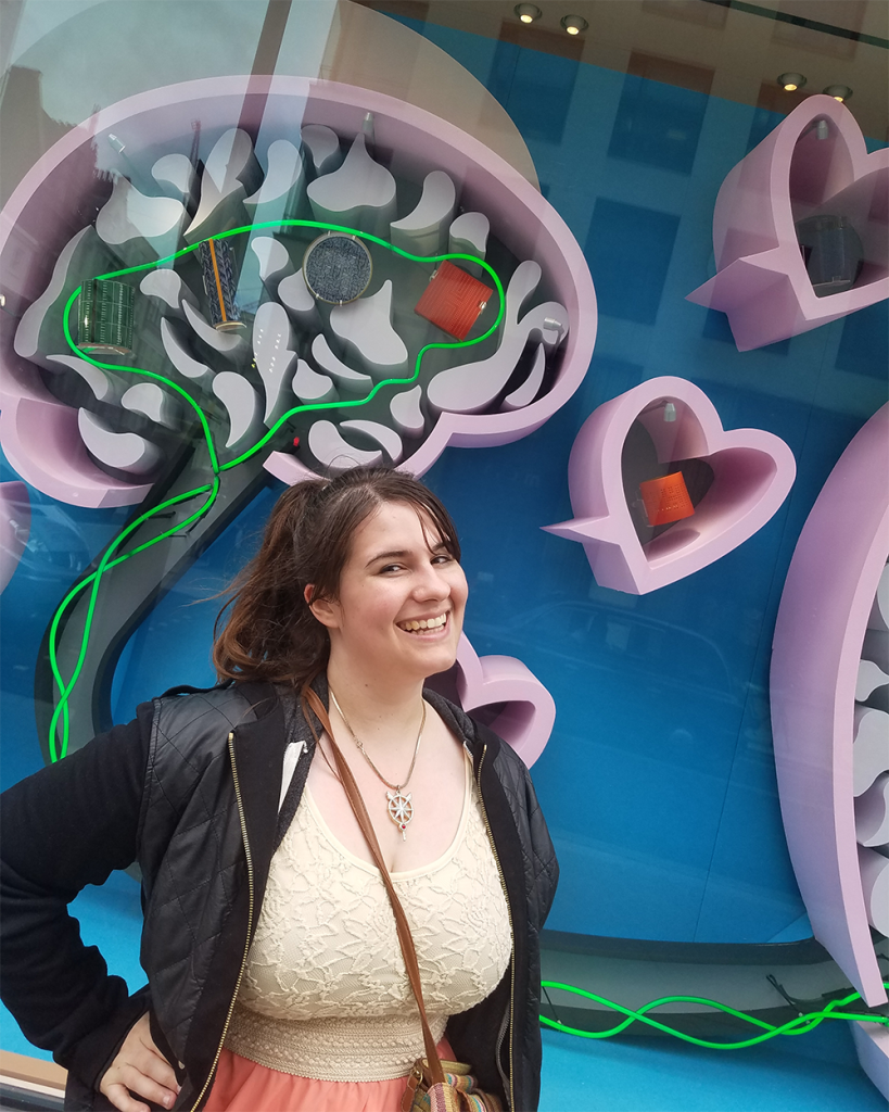 Image of Briana in a dress and blazer outside of a storefront display, which shows an artistic rendering of a brain above her head and a dialogue bubble shaped like a heart to her side. 