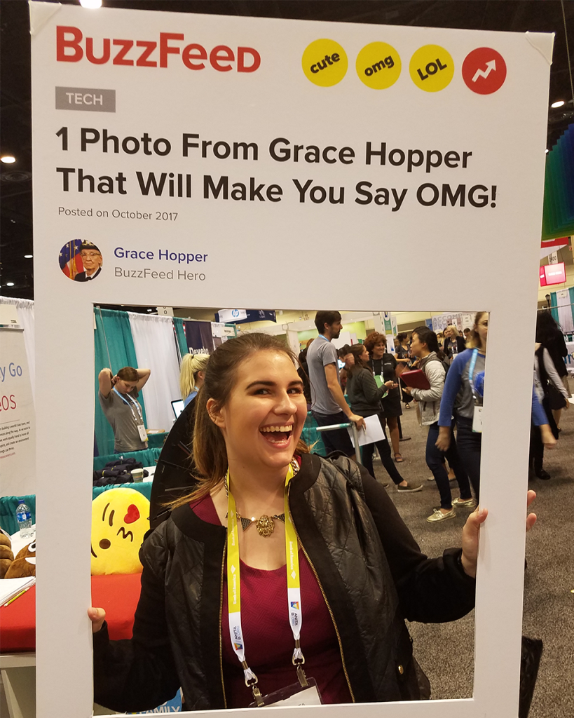 Image of Briana in a blazer smiling from a cardboard cutout she is holding up. The cutout is designed to look like a Buzzfeed article, with a title that says "1 Photo from Grace Hopper That Will Make You Say OMG!"